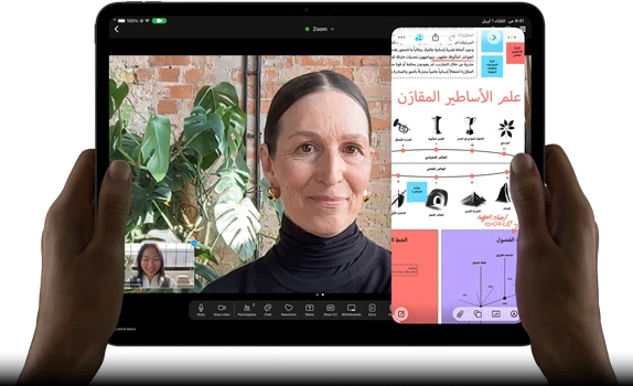 جهاز iPad Pro، الجزء الأمامي الخارجي، الوضع الأفقي، لون أسود فلكي، زوايا مدورة، إطار شاشة أسود، يدان تحملان الجهاز من الجانب الأيسر والأيمن، شاشة تعرض محادثة عبر فيس تايم تتم ترجمتها مباشرة، وتطبيق تعليمي يُظهر عرضاً تقديمياً بعنوان 'علم الأساطير المقارَن' مع نصوص ومخططات وملاحظات مكتوبة بخط اليد