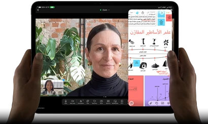 جهاز iPad Pro، الجزء الأمامي الخارجي، الوضع الأفقي، لون أسود فلكي، زوايا مدورة، إطار شاشة أسود، يدان تحملان الجهاز من الجانب الأيسر والأيمن، شاشة تعرض محادثة عبر فيس تايم تتم ترجمتها مباشرة، وتطبيق تعليمي يُظهر عرضاً تقديمياً بعنوان 'علم الأساطير المقارَن' مع نصوص ومخططات وملاحظات مكتوبة بخط اليد