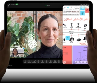جهاز iPad Pro، الجزء الأمامي الخارجي، الوضع الأفقي، لون أسود فلكي، زوايا مدورة، إطار شاشة أسود، يدان تحملان الجهاز من الجانب الأيسر والأيمن، شاشة تعرض محادثة عبر فيس تايم تتم ترجمتها مباشرة، وتطبيق تعليمي يُظهر عرضاً تقديمياً بعنوان 'علم الأساطير المقارَن' مع نصوص ومخططات وملاحظات مكتوبة بخط اليد
