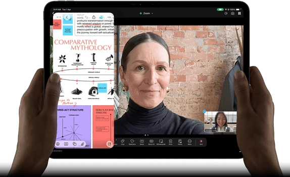 iPad Pro, front exterior, landscape orientation, space black color, rounded corners, black display bezel, hands holding left and right sides, display showing a FaceTime conversation being live translated, and an educational app showing a presentation of "Comparative Mythology" with text, diagrams, and handwritten notes