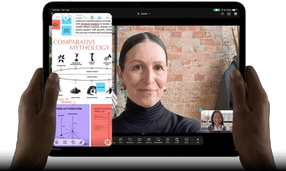 iPad Pro, front exterior, landscape orientation, space black color, rounded corners, black display bezel, hands holding left and right sides, display showing a FaceTime conversation being live translated, and an educational app showing a presentation of "Comparative Mythology" with text, diagrams, and handwritten notes