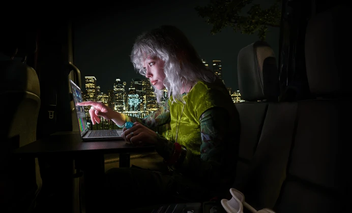 شخص يجلس في المقعد الخلفي لسيارة فان، باب الراكب المفتوح يكشف عن أفق مدينة في الليل، جهاز MacBook Pro مع شاشة PROLED XDR موضوع على طاولة عمل، ضوء الشاشة ينعكس على الشخص أثناء استخدامه لميزة اللمس من أجل تعديل فيديو في برنامج DaVinci Resolve Studio‏