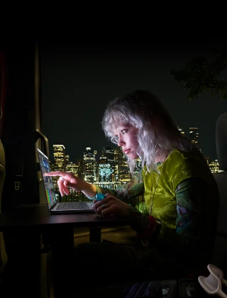شخص يجلس في المقعد الخلفي لسيارة فان، باب الراكب المفتوح يكشف عن أفق مدينة في الليل، جهاز MacBook Pro مع شاشة PROLED XDR موضوع على طاولة عمل، ضوء الشاشة ينعكس على الشخص أثناء استخدامه لميزة اللمس من أجل تعديل فيديو في برنامج DaVinci Resolve Studio‏