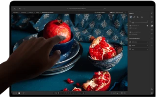 MacBook Pro with PROLED XDR Display with a still life image of pomegranates with blue ceramics and textiles, a hand reaches toward the screen to interact with the image