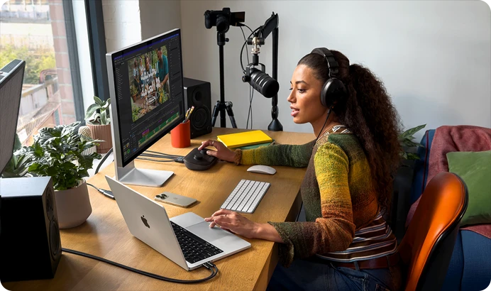 امرأة في منزلها تعمل خلف مكتبها على MacBook Pro متصل بشاشتين خارجيتين وترتدي سماعات رأس وتتحدث في الميكروفون
