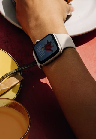 ذراع شخص يتناول الفطور وعلى معصمه Apple Watch SE 3 بإطار من الألومنيوم ولون ضوء النجوم، وشاشة بميزة "التشغيل دوماً"، وواجهة الساعة تحمل صورة بورتريه، وحزام رياضي بلون ضوء النجوم